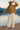 Woman in oatmeal crochet knit cardigan, cream pants, beige espadrilles and hat, standing on dock, mountain view