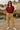 Woman in a maroon shirt and beige pants standing by a waterfront with buildings in the background