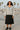 Woman in heather gray jacquard zip jacket, pleated skirt, black sneakers, holding skateboard outdoors