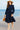 Woman in navy ruffle shoulder midi dress walking seaside promenade with straw hat