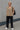 Standing White blonde woman in beige vest and black trousers with white sneakers, gray wall, diffused light