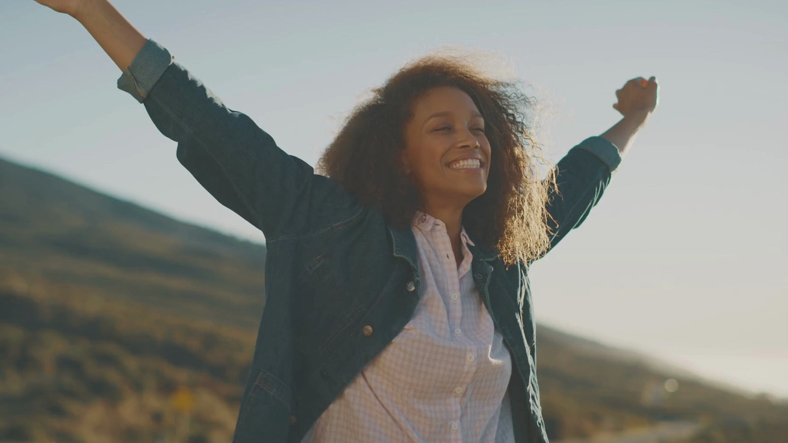 A joyful woman with curly hair, arms raised, wearing a denim jacket and a light checkered shirt, embodies the carefree spirit of the Women's Boutique.