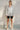 Woman in satin shirt with black bike shorts and white heels posing by white wall