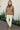 Woman wearing a brown sweater and white pants standing on a paved path with flowers in the background