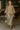 Model walking in taupe textured knit V-neck top with patterned skirt and sandals, modern cafe interior.