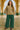 Woman posing in taupe textured knit V-neck top with green wide-leg pants, seaside archway.
