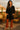 Woman in a black dress and orange pants standing on a wooden deck with cacti and chairs in the background.